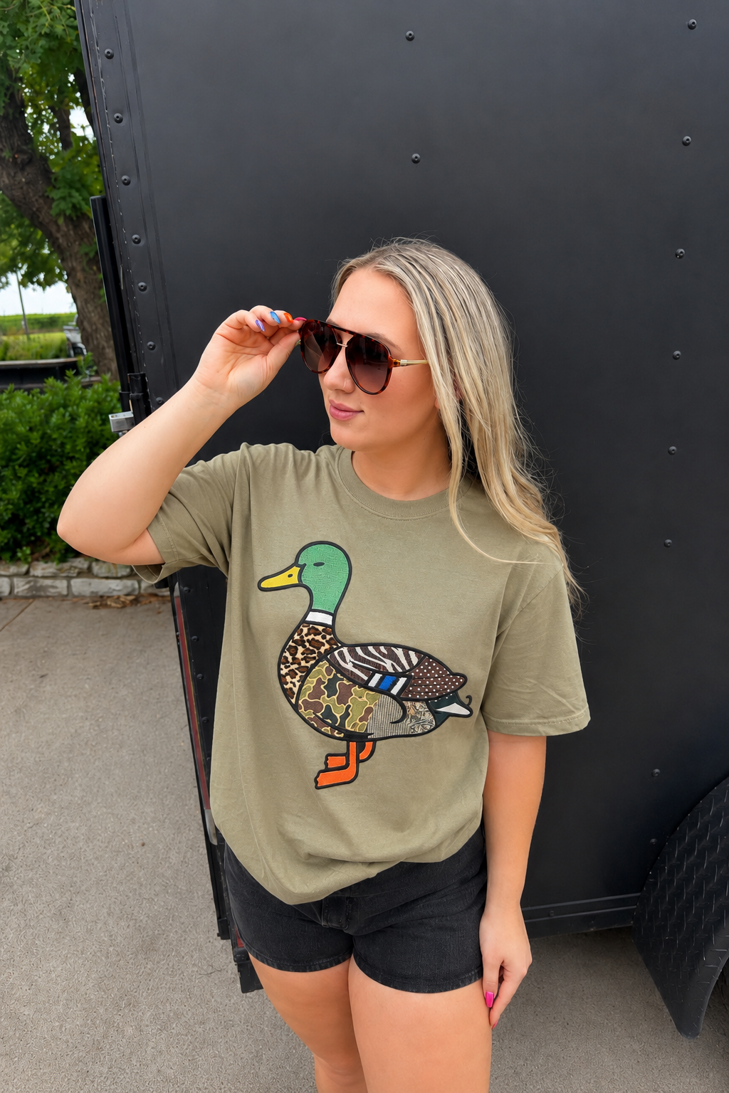 A woman wearing a muted olive green graphic t-shirt with a detailed mallard duck print, paired with black shorts, standing outdoors next to a black trailer.