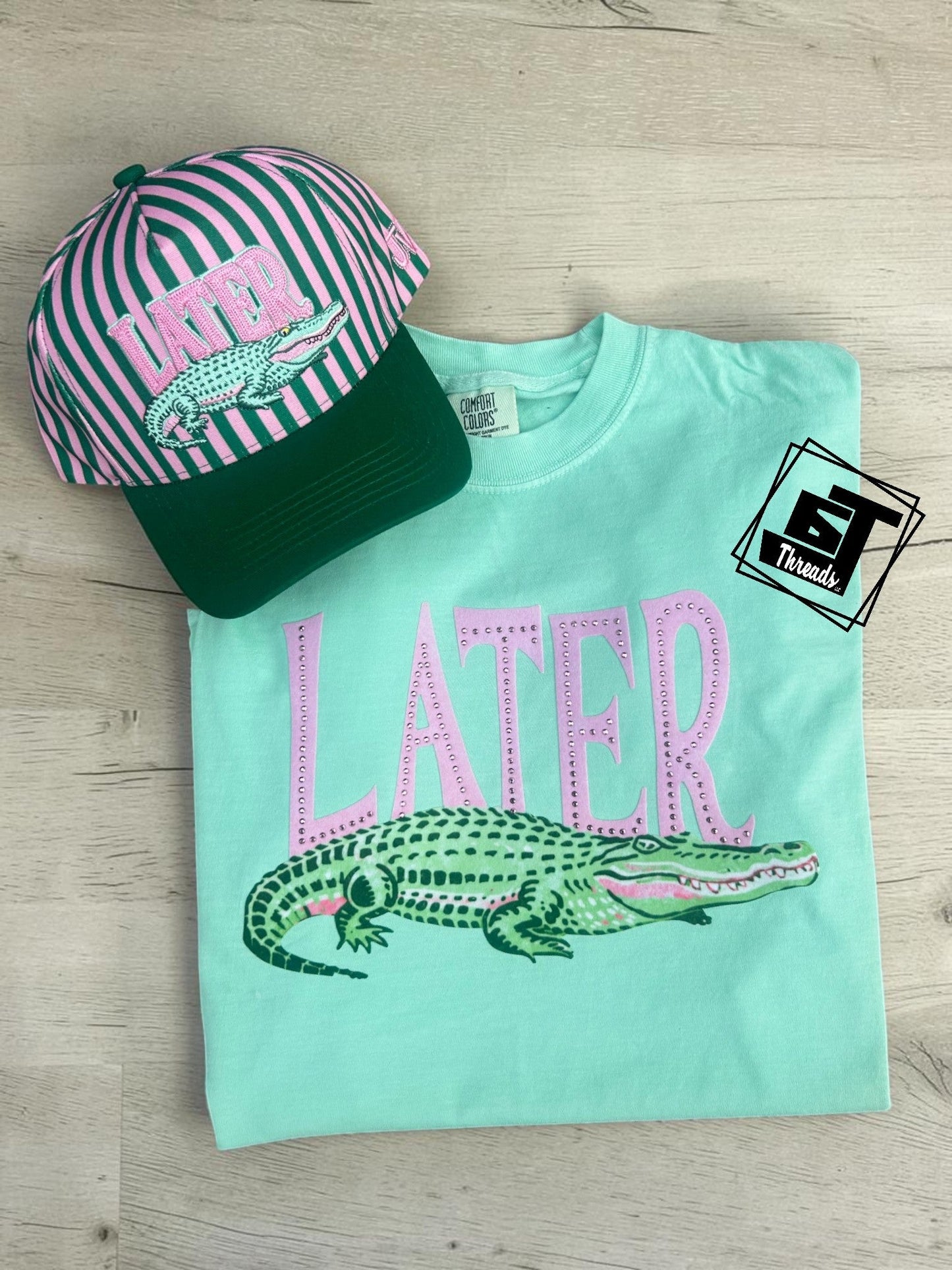Mint green t-shirt with alligator graphic and 'LATER' text, paired with a striped cap on a wooden surface.