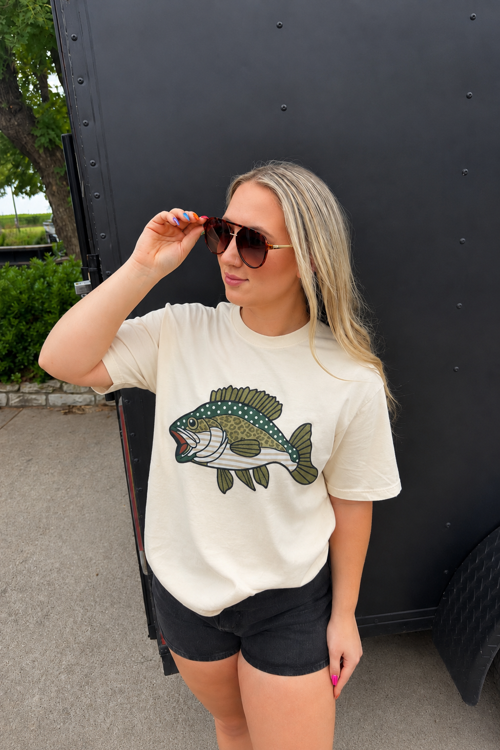 A woman wearing a cream-colored t-shirt with a green bass fish graphic, paired with black shorts, standing in front of a black trailer.