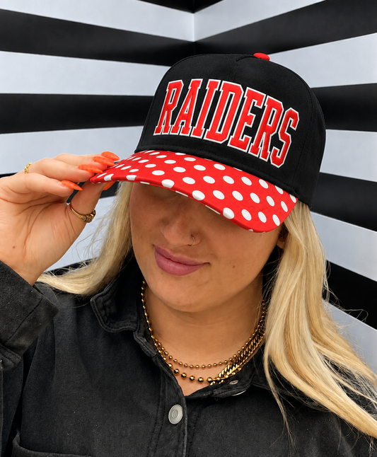 A woman wearing a black baseball cap with 'RAIDERS' in red letters and a red brim covered in white polka dots, posing against a black and white striped background.