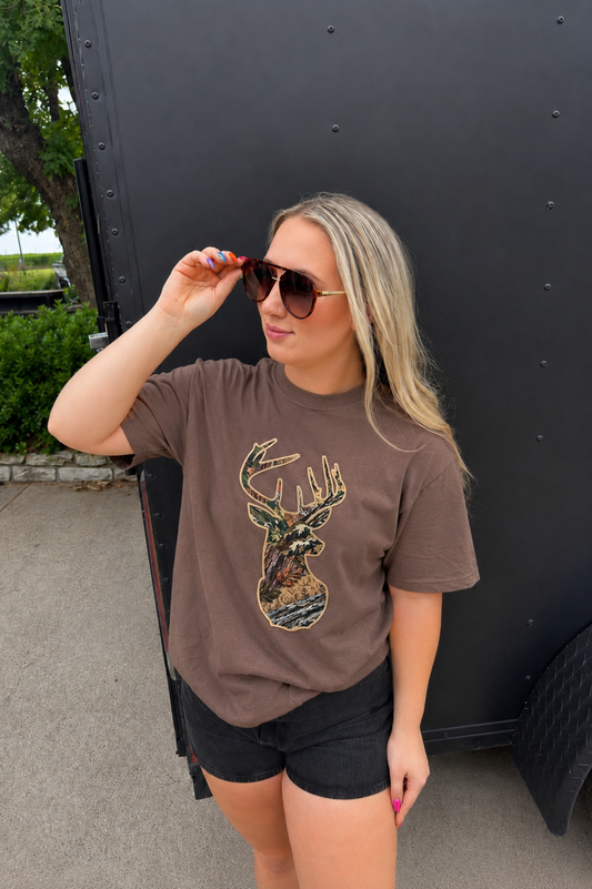 A woman wearing a brown graphic t-shirt with a sequined deer head design, black shorts, and sunglasses, standing outdoors in front of a black trailer.