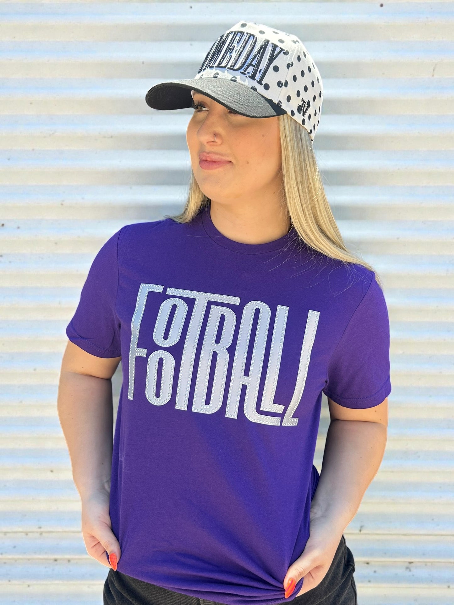 Woman wearing a purple 'FOOTBALL' t-shirt and a cap with a polka dot pattern against a corrugated metal background.