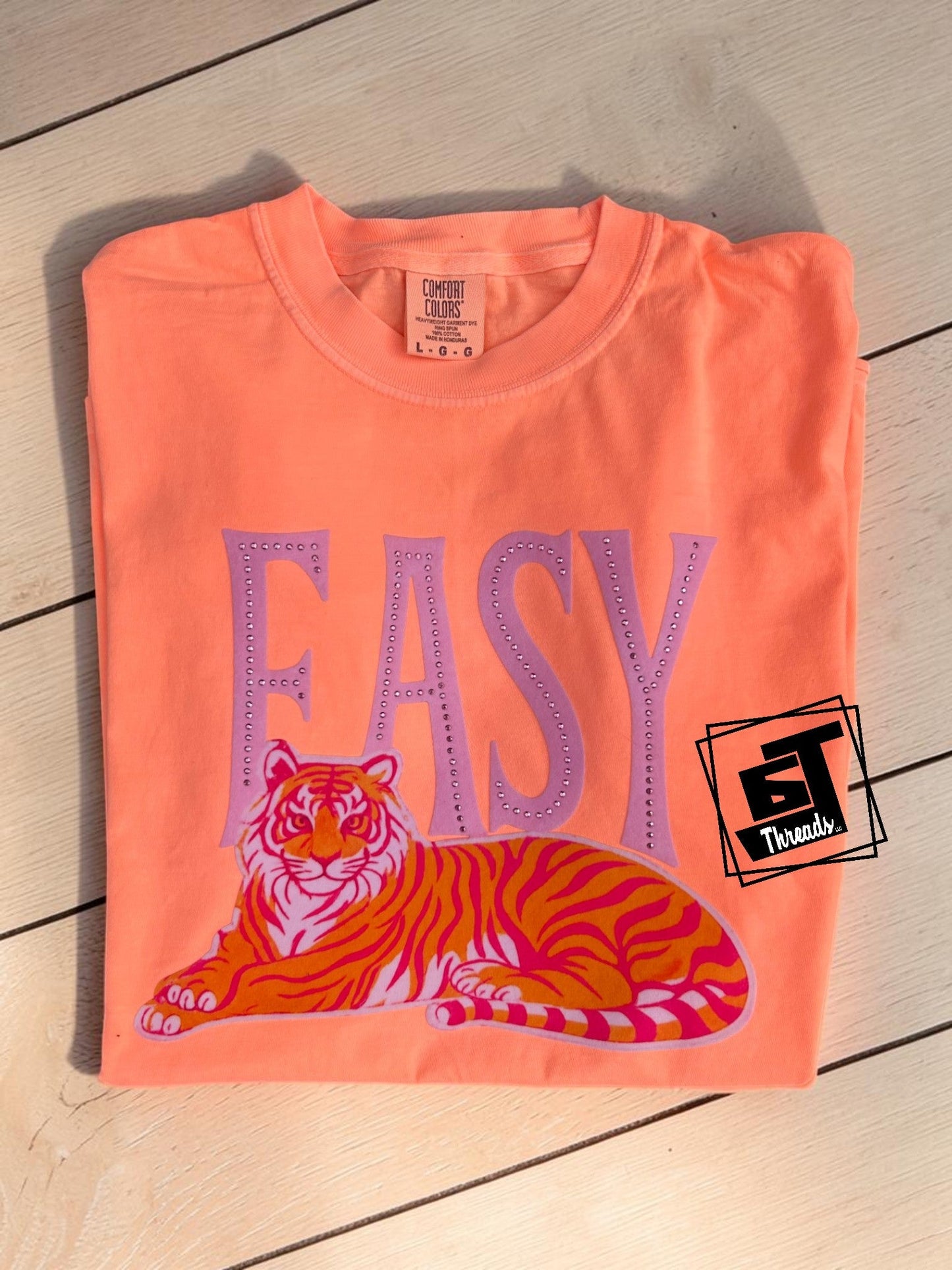 Folded orange t-shirt with a tiger graphic and 'EASY' text on a wooden surface