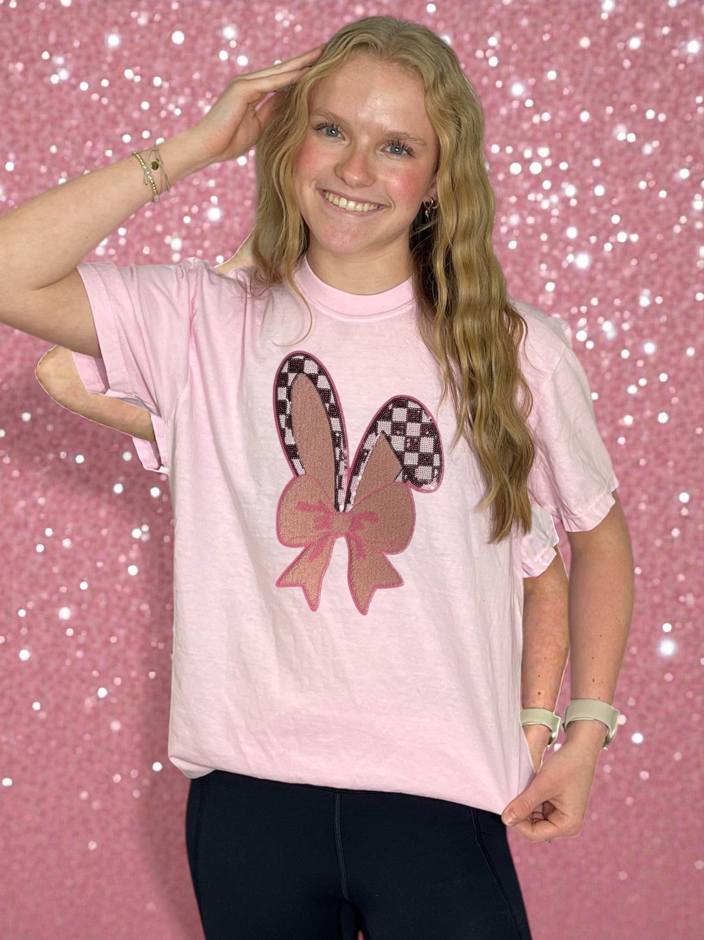 Person wearing a pink t-shirt with a checkered bow design on a pink glittery background