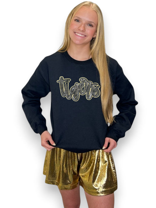 Person wearing a black long-sleeve shirt with 'Tigers' printed on it and gold sequin shorts.
