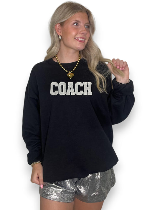 Woman wearing a black sweatshirt with 'COACH' text and silver shorts on a white background