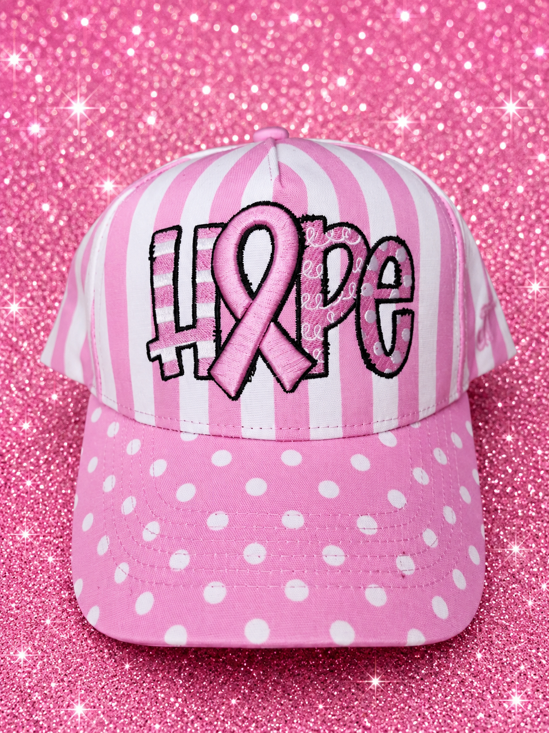 A pink and white striped baseball cap with a pink polka-dot brim, featuring the word 'HOPE' in stylized letters where the 'O' is a pink awareness ribbon. The cap is set against a sparkly pink background.