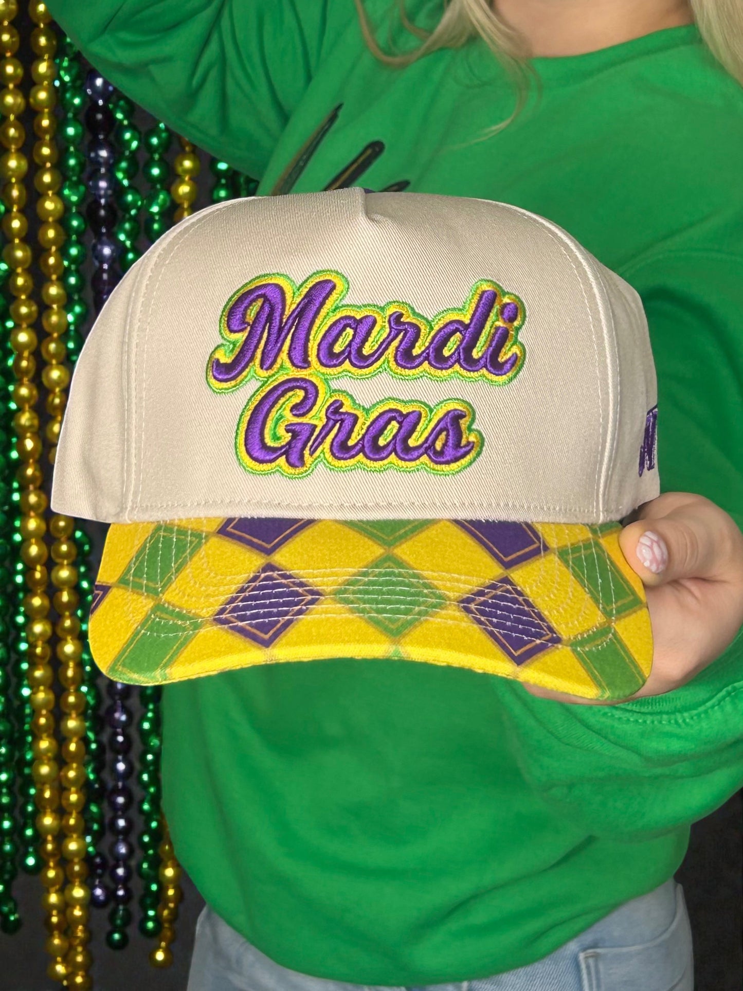 Beige cap with 'Mardi Gras' text held by a person wearing a green shirt, with Mardi Gras beads in the background.