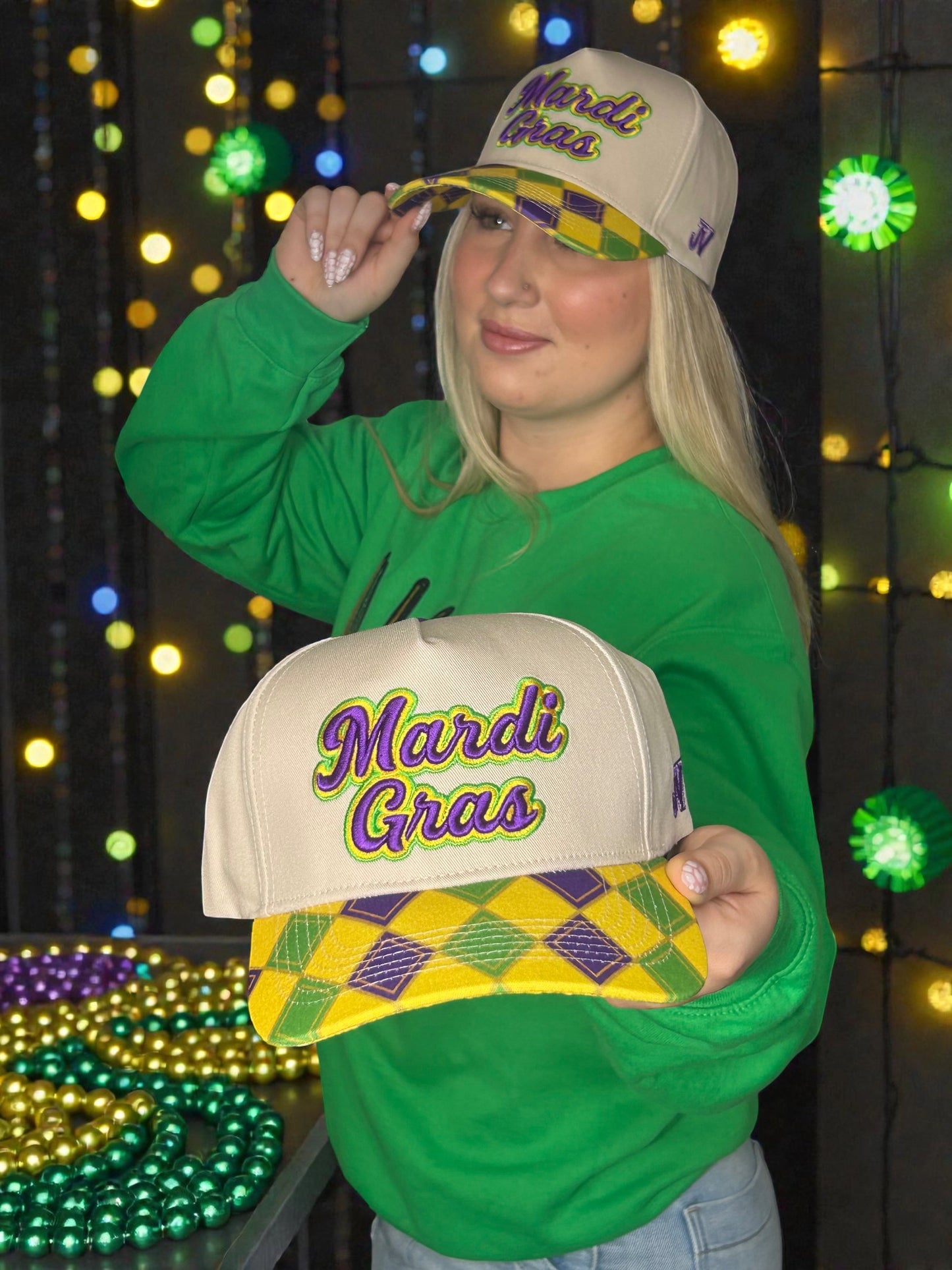 Person wearing a green shirt and holding two Mardi Gras-themed hats against a festive background with lights.