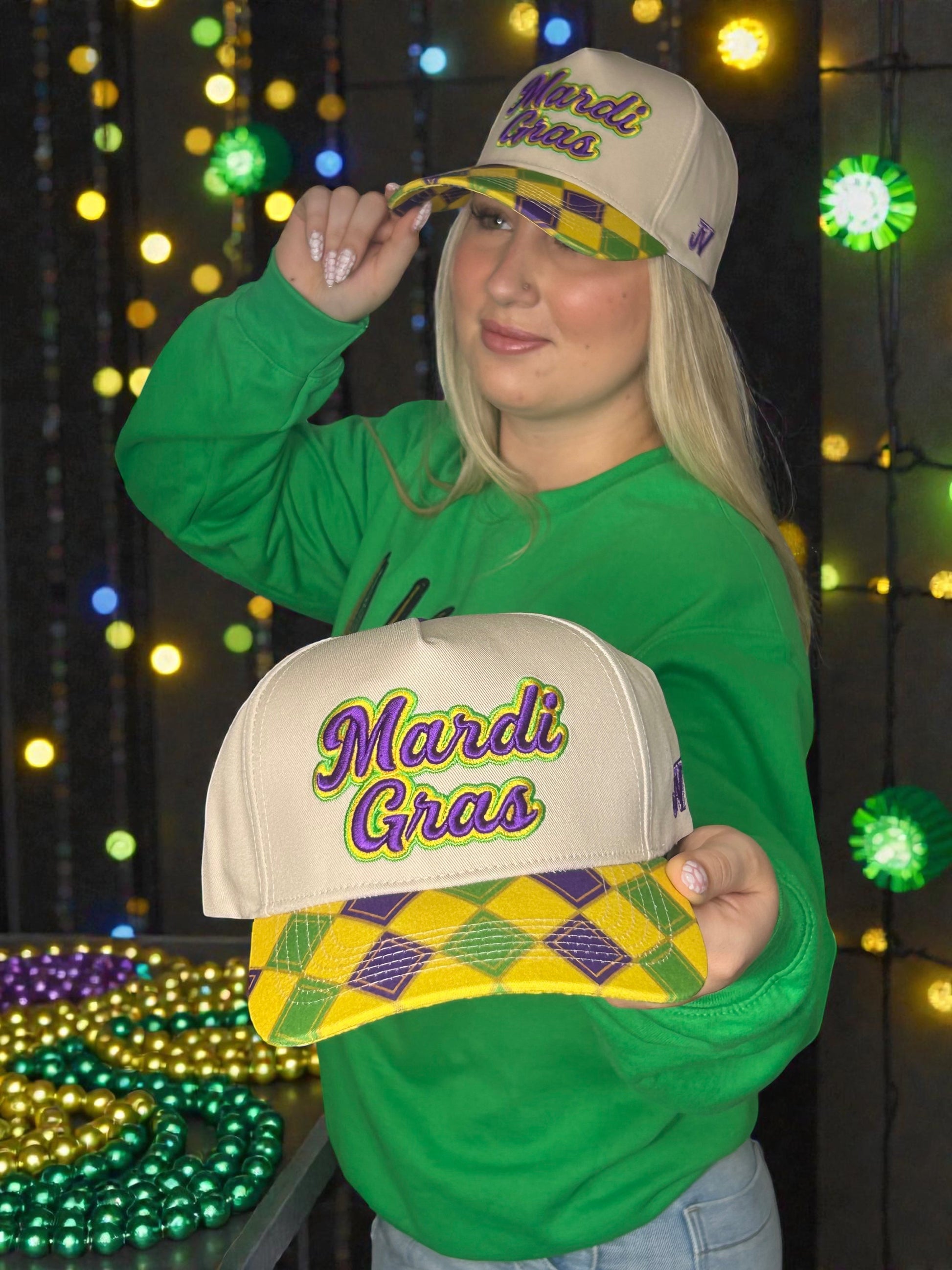 Person wearing a green shirt and holding two Mardi Gras-themed hats against a festive background with lights.