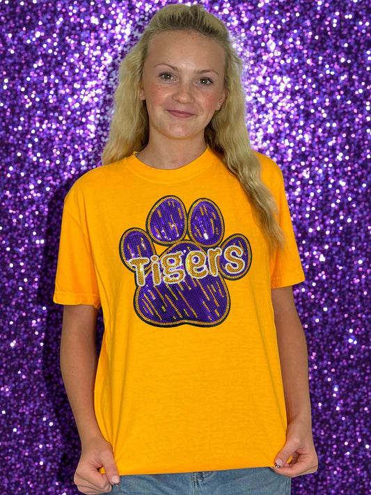 Person wearing a yellow t-shirt with a purple paw print and 'Tigers' text against a purple glittery background